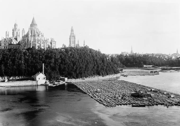 Black and white photograph of nineteenth-century Ottawa. The foreground shows a landing point with timber rafts; grand parliamentary buildings are in the background, a belt of dark trees separates the water from the buildings.