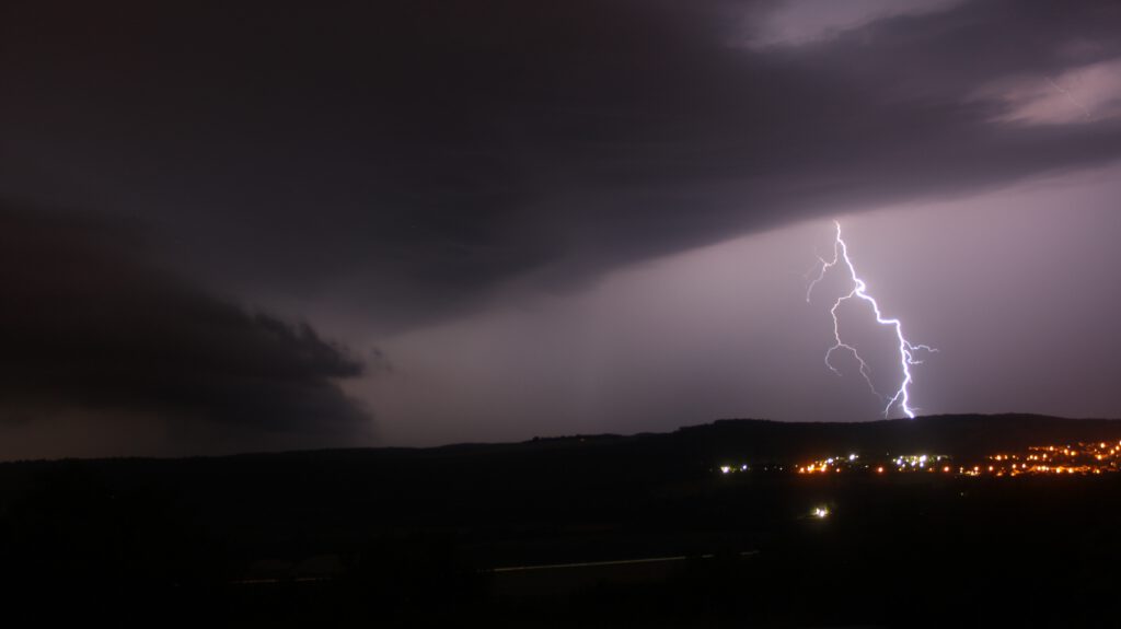 Lightning striking a hill in a dark landscape. The lights of a settlement can be seen in the right of the image, below the hill where the lightning strikes