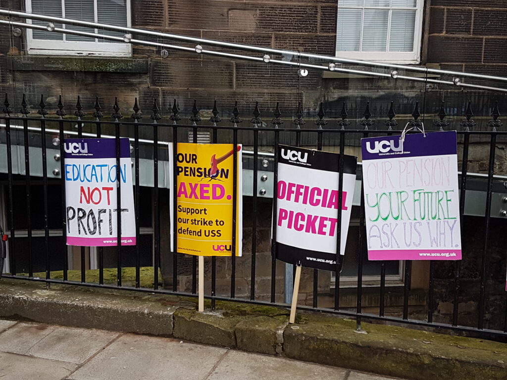 Four placards against a black fence, reading (from left to right): "Education not profit" - "Our pension AXED. Support our strike to defend USS" - "Official picket" - "Our pension your future, ask us why"