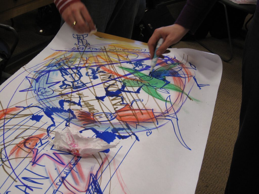 A large sheet of paper that (at least) two people have been drawing on using brightly colored felt-tip pens. At this moment we can see one hand drawing using a blue pen, while a second person's hand clutches a piece of tissue paper that they seem to be using for blotting. The drawing produced so far is a confused mix of different figures, colors and shapes, with many lines across it and large circles linking many of the figures together. 'I CAN DRAW' has been written in different ways across various parts of the image.